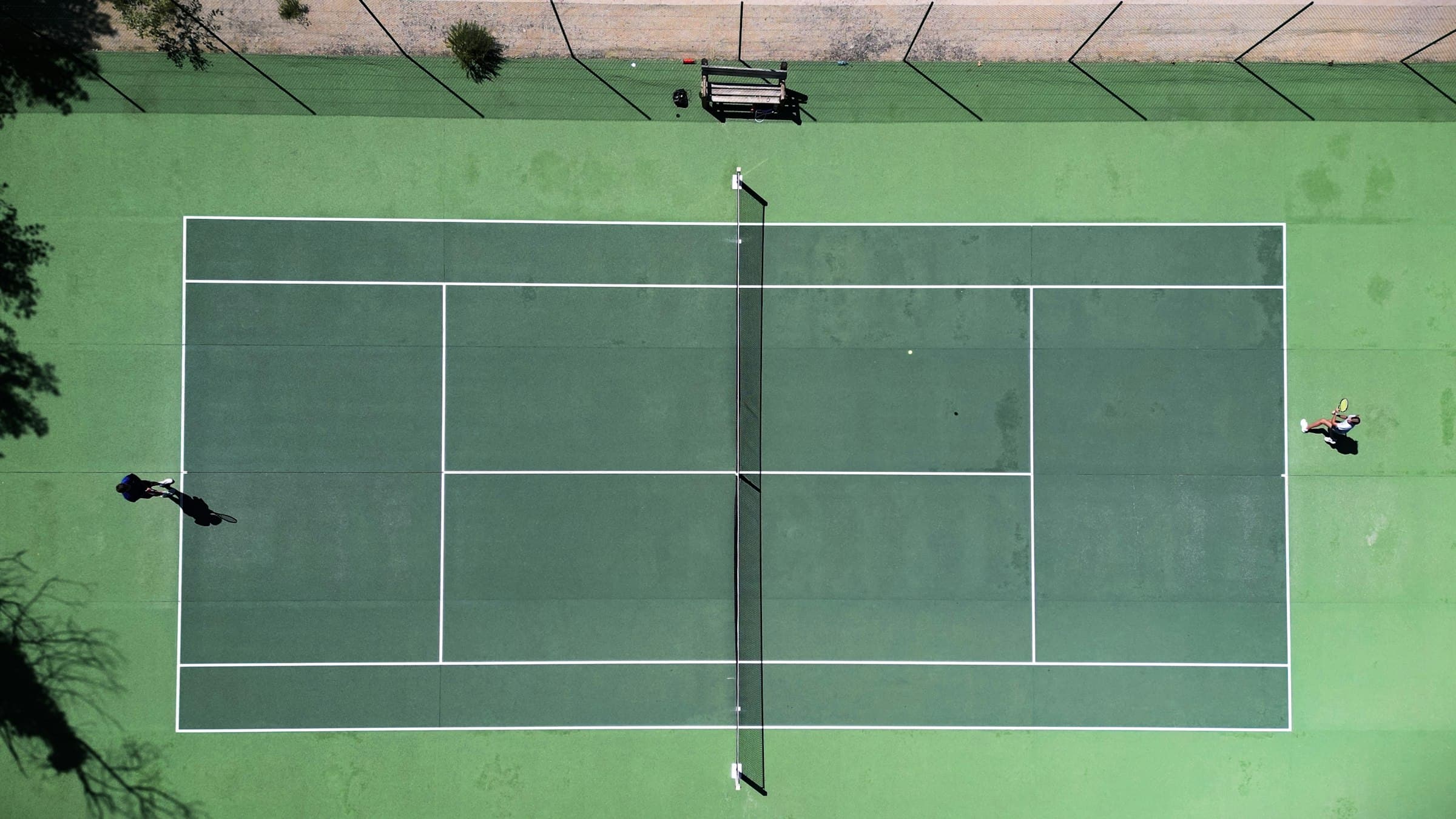 A hard court seen from directly above, late in the day: two players mid-rally, the ball hanging just past the net, their shadows stretched across the tramlines.