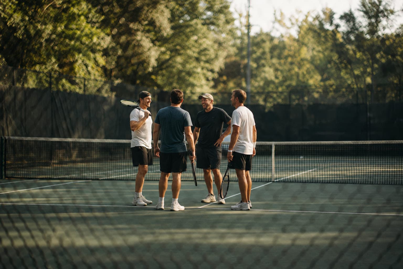Four players standing together at the net after a match on a tree-lined hard court, rackets down, mid-conversation in late afternoon light.